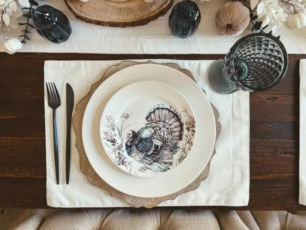 A thanksgiving placesetting that has a blue and white turkey plate, black flatware and a blue goblet