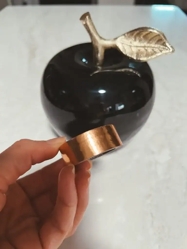 A hand holding a roll of copper washi tape in front of a black apple on a counter
