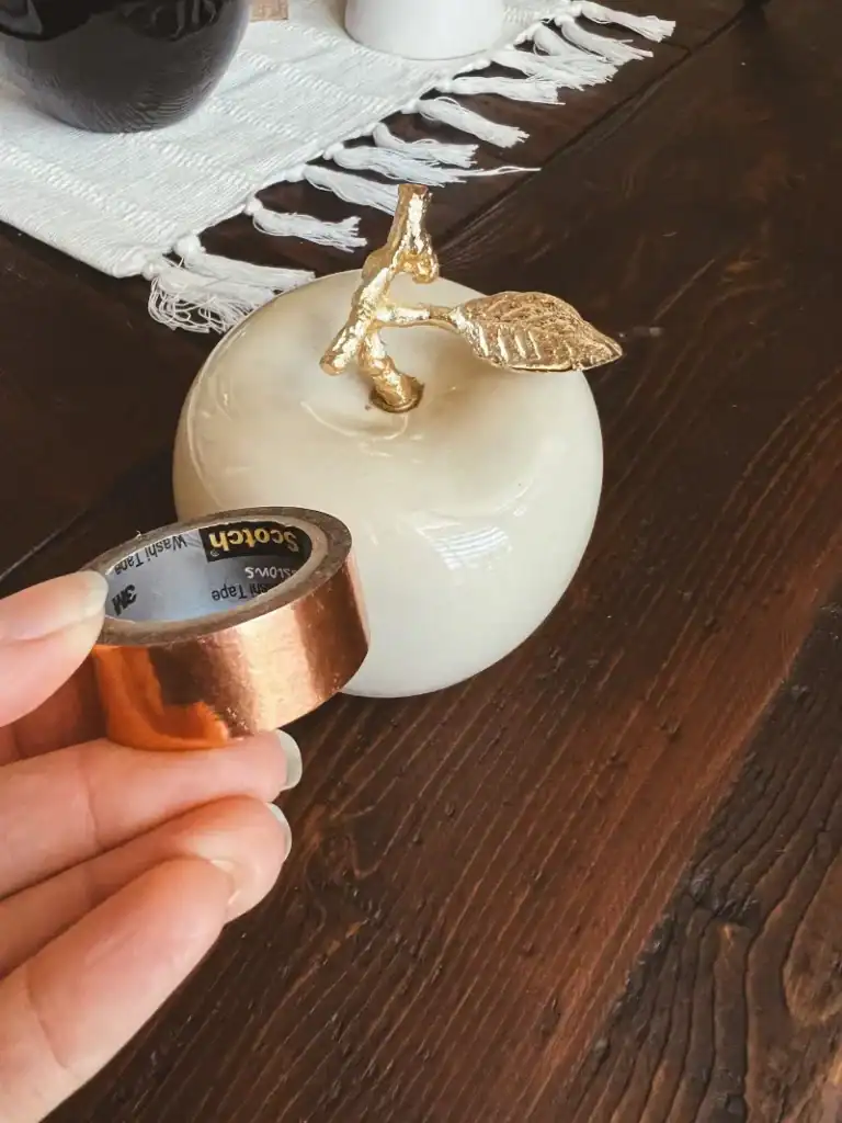 A ceramic white apple with a white hand holding a roll of copper colored washi tape in front of it
