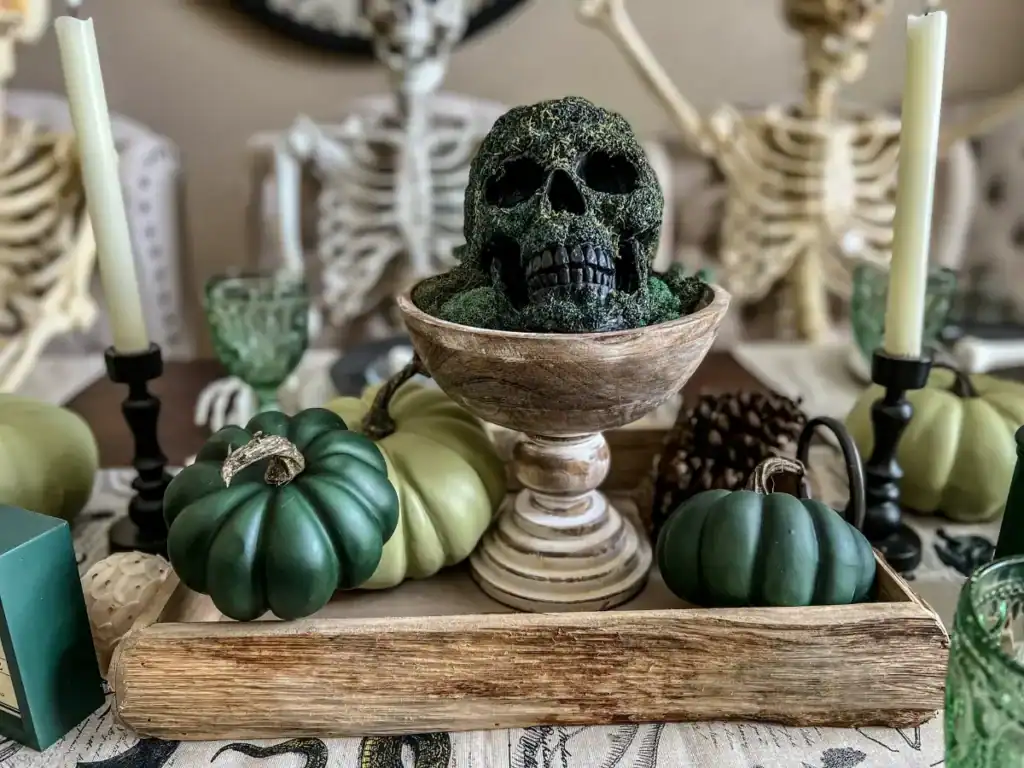 A skull covered in green moss on a pedestal for a halloween tablescape centerpiece placed in a tray with green pumpkins around it