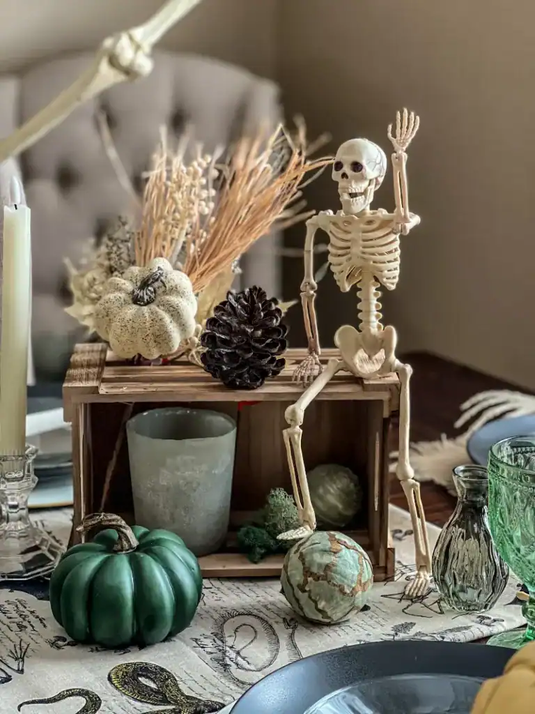 A skeleton waving sitting on top of a crate next to a pinecone and pumpkins 