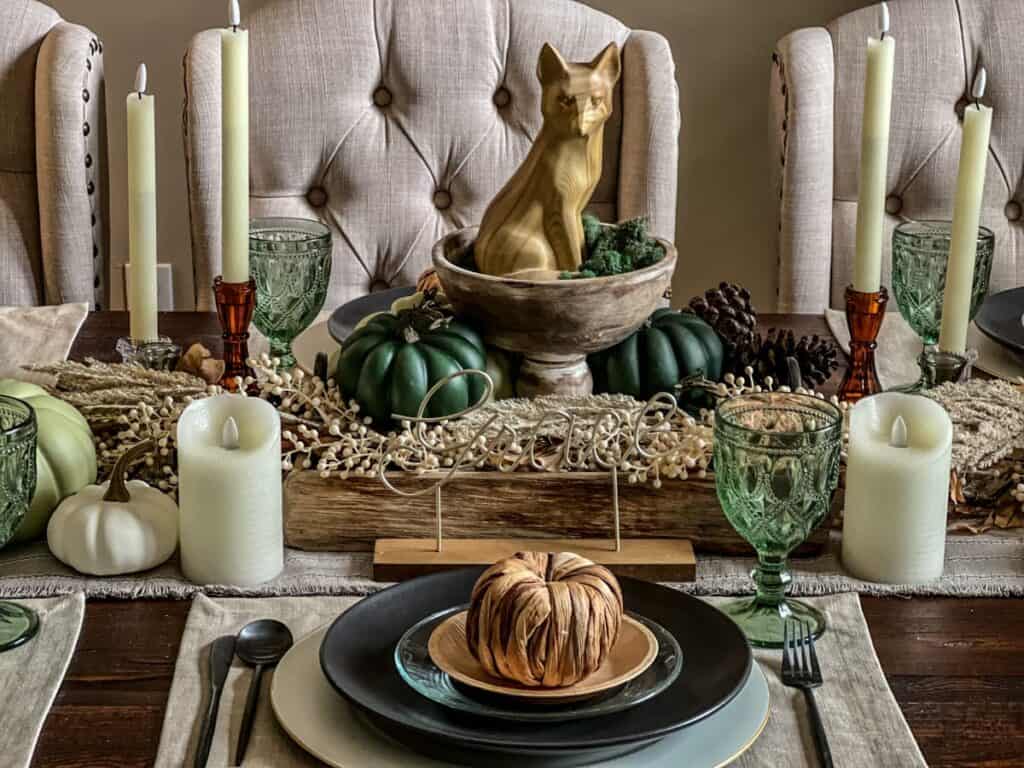 A Thanksgiving tablescape with a fox centerpiece, green decor, a black plate and raffia pumpkin on plate